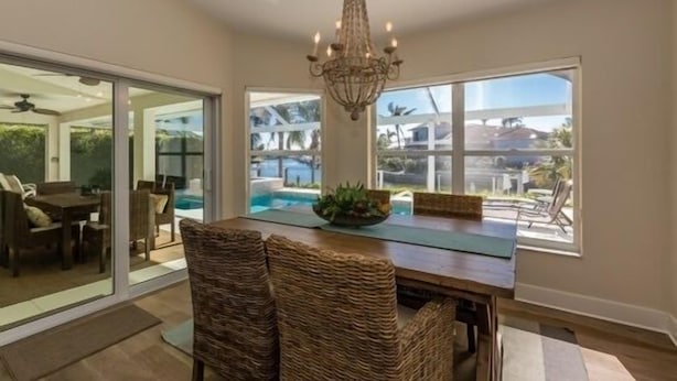 Uma sala de jantar com uma mesa grande, cadeiras de vime, um candelabro e grandes janelas com vista para uma piscina e área de estar ao ar livre.