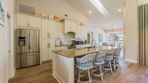 A modern kitchen with white cabinetry, a stainless steel refrigerator, a central island with bar stools, and a large window providing natural light. - Perfect 4/2  home for your Marco Island vacations (Marco Island)