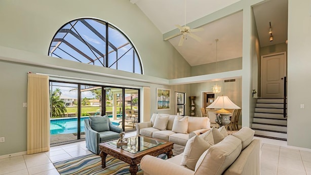 A spacious living room with a large window, a ceiling fan, and a view of a swimming pool. - 4 bedroom, 3 bath home with heated pool (Marco Island)