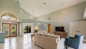 A spacious living room with a large sofa, a blue armchair, and a television. There is a door leading to a balcony with plants. - 4 bedroom, 3 bath home with heated pool (Marco Island)