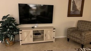 Un salon avec un téléviseur à écran plat fixé au mur, un meuble de rangement en bois en dessous, un fauteuil à motifs et une grande plante en pot.