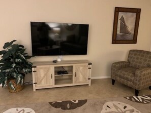 A living room with a flat-screen TV mounted on the wall, a wooden entertainment unit below, a patterned armchair, and a large potted plant.