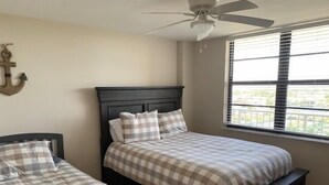 Une chambre avec deux lits, un ventilateur de plafond, une fenêtre avec des stores et un support mural.
