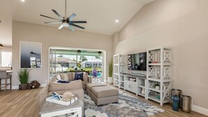 A modern living room with a sectional sofa, ottoman, and a ceiling fan. There is a TV on a white shelving unit and a large window with a view of the outdoors. - Beautiful 3Bdr Home located in the heart of Marco. (Marco Island)