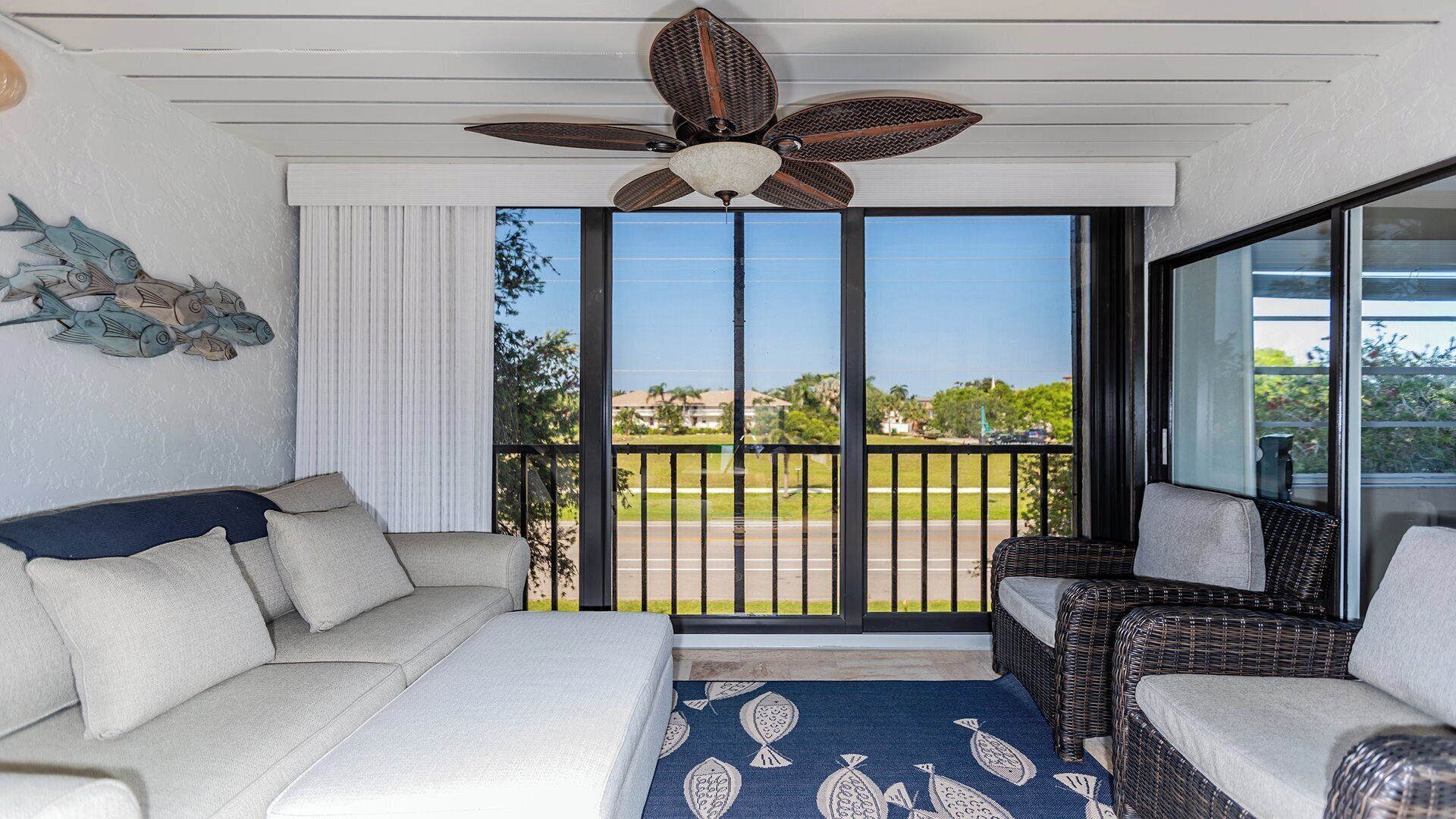 Habitación con sofá blanco, una silla de mimbre, ventilador de techo y vistas a un paisaje verde a través de un ventanal corredero.