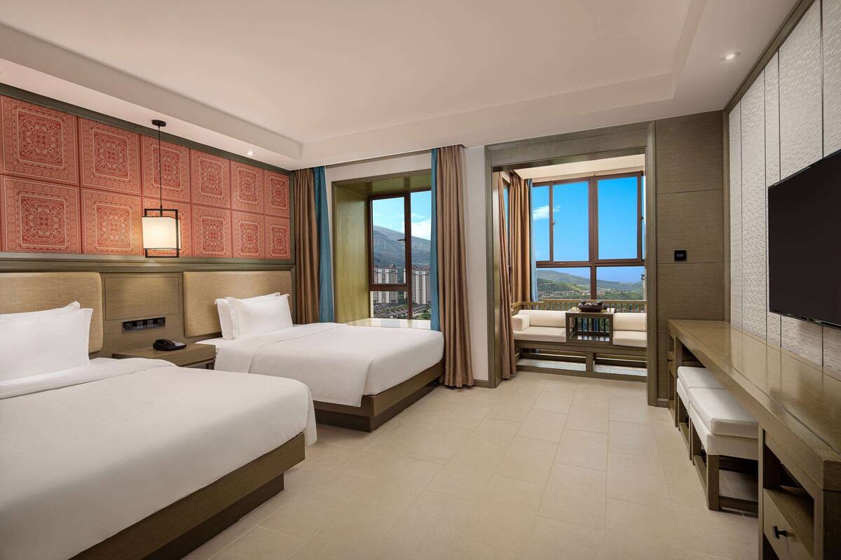 Ein Hotelzimmer mit zwei Betten, einem Schreibtisch, einem Fernseher und einem Balkon mit Blick auf die Stadt.
