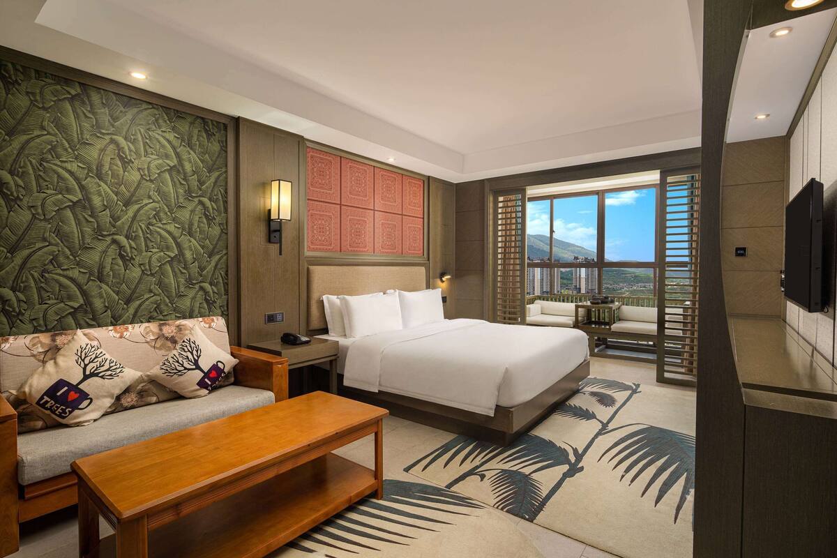 Ein Hotelzimmer mit einem großen Bett, einer Couch, einem Holztisch und Blick auf die Stadt durch große Fenster.