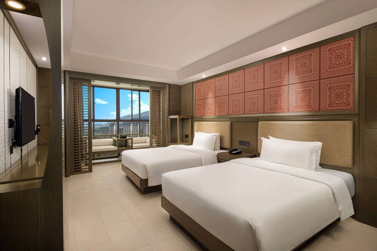 Ein Hotelzimmer mit zwei Betten, einem Fernseher, einem Balkon mit Blick auf die Stadt und einer Zierleiste.