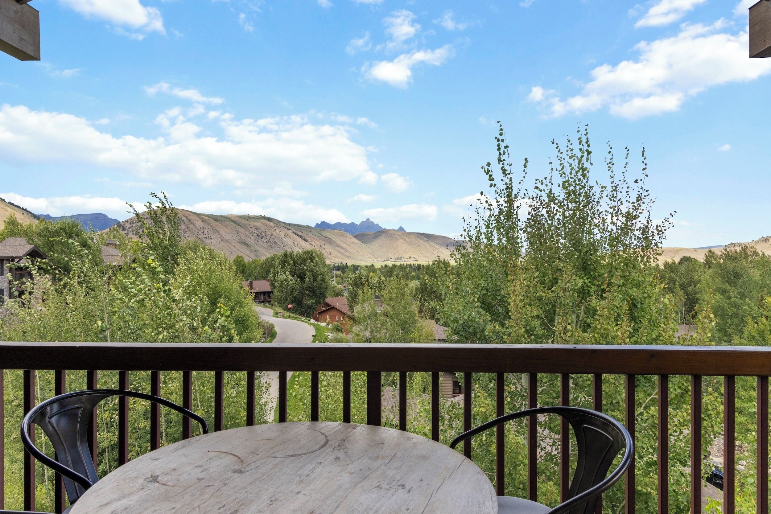 A balcony with a wooden table and metal chairs, overlooking a mountainous landscape with greenery and a clear sky.