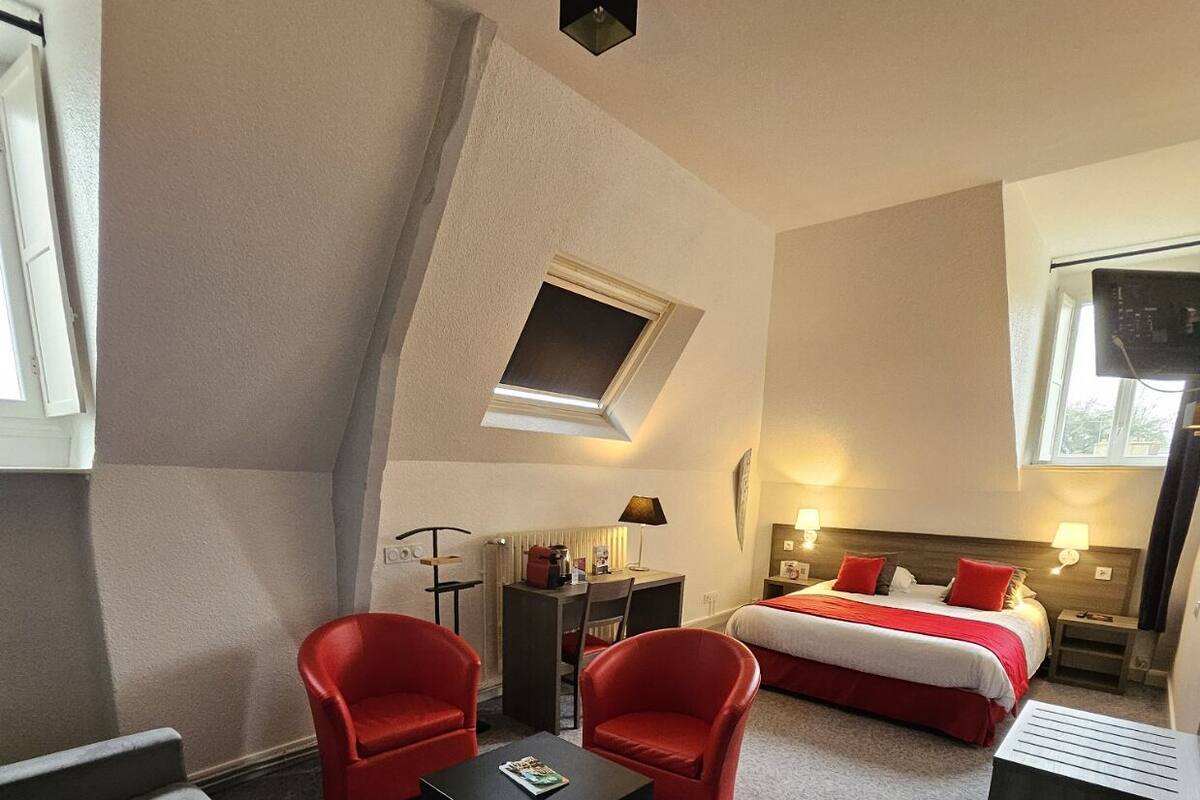 A hotel room with a bed, two red chairs, a small table, a desk with a lamp, and a skylight.