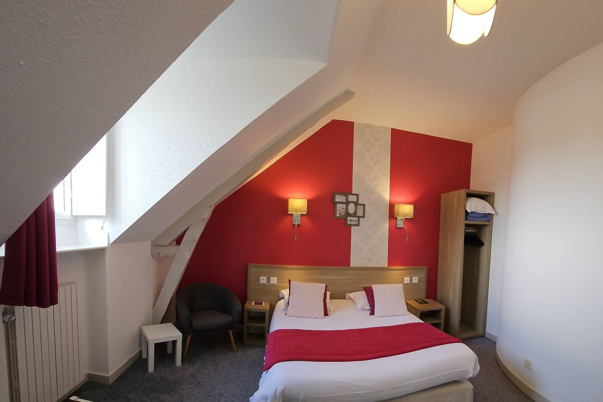 A hotel room with a sloped ceiling, a bed with white bedding and a red accent, two bedside tables with lamps, a chair, and a small desk.