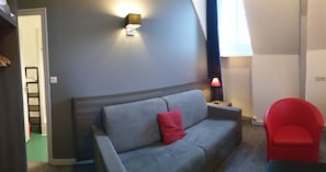 Una habitación con un sofá gris, un sillón rojo, una mesita con una lámpara y una lámpara de pared.