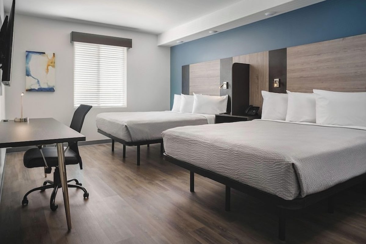 A hotel room with two single beds, a desk with a chair, a flat-screen TV, and a window with blinds.