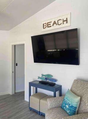 A living room with a flat-screen TV mounted on the wall, a small table with a decorative item, a couch with a beach-themed pillow, and a door leading to another room.