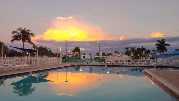 Ein Swimmingpool mit Liegestühlen und Palmen, umgeben von Gebäuden und einem Sonnenuntergangshimmel.