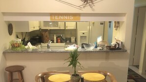 A dining area with a wooden table and chairs, a kitchen in the background, and a tennis court sign on the wall. - Ultimate Tennis Condo at World Tennis Center (Naples)