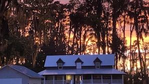 Un edificio de dos pisos con un porche y un garaje, rodeado de árboles y con el atardecer de fondo.