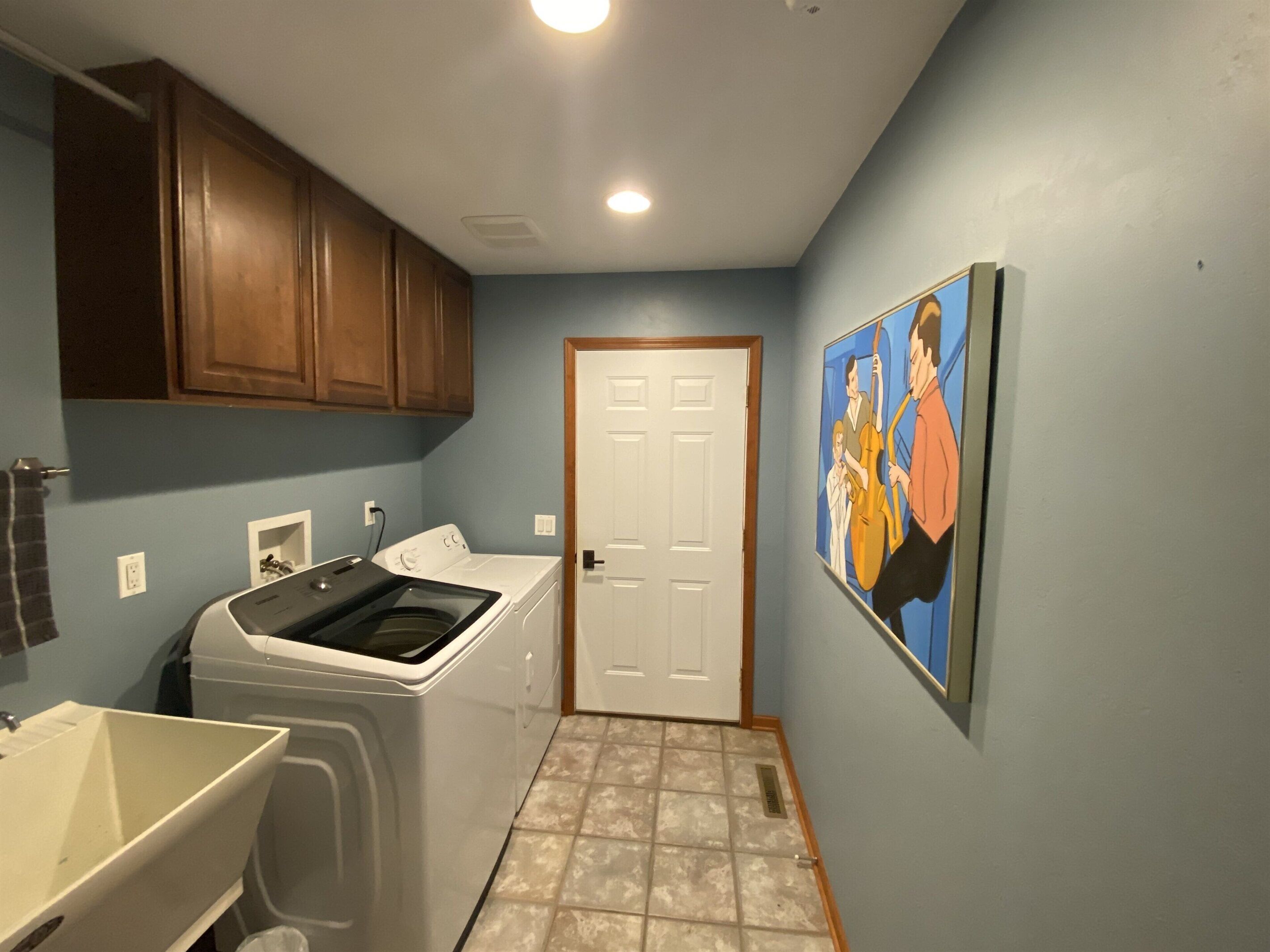 Un lavadero con lavadora, secadora y lavabo. Hay una puerta que da a otra habitación y un cuadro en la pared.