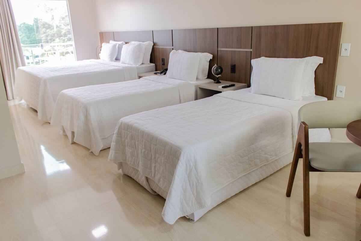 A hotel room with three single beds, a wooden headboard, a bedside table with a lamp, and a chair.
