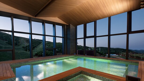 A modern indoor swimming pool with large windows offering a view of a hilly landscape.