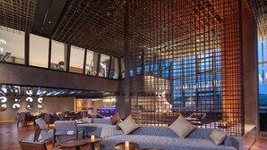 A modern lounge area with seating and tables, a decorative wall with a grid pattern, and a view of the cityscape through large windows.