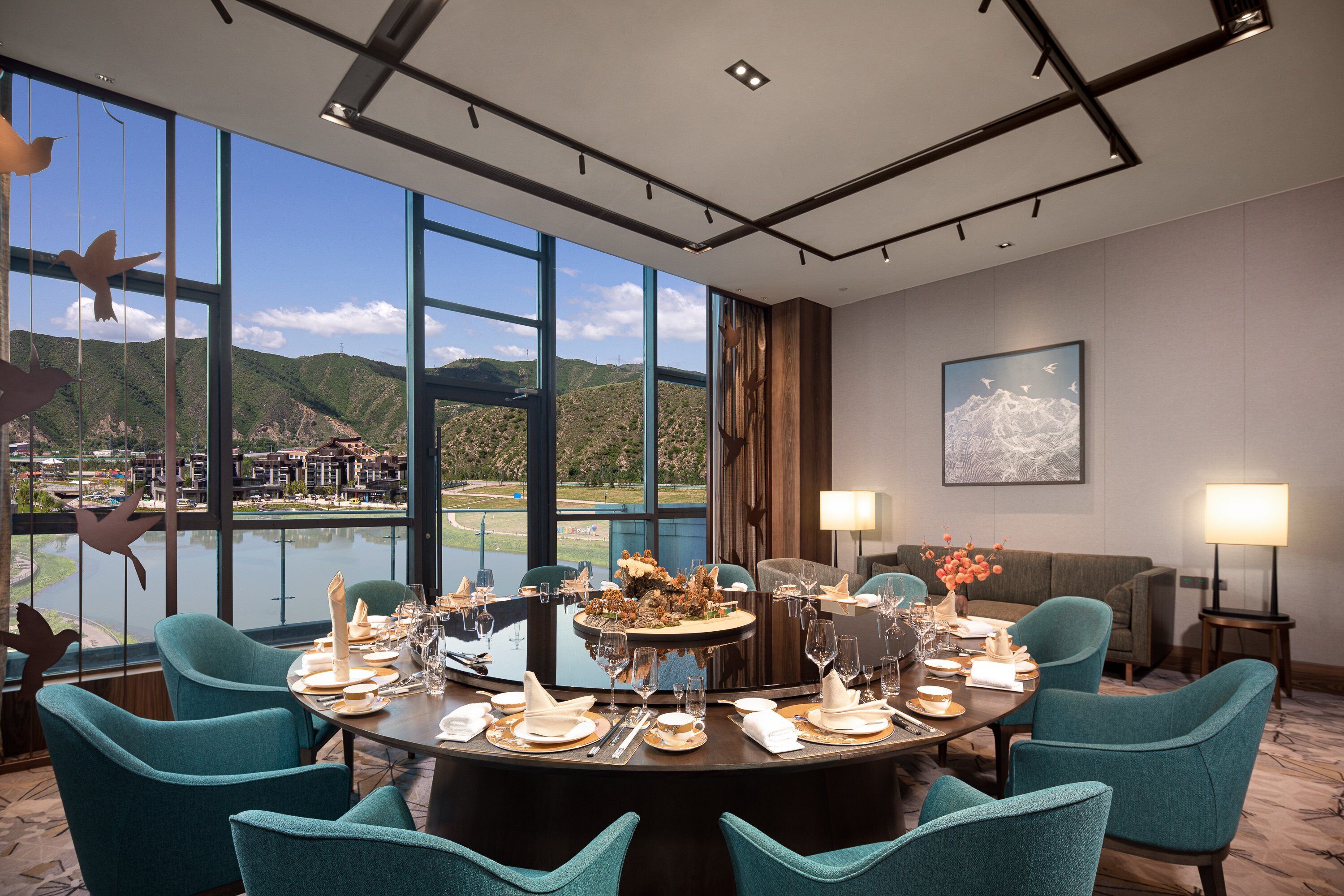 A dining area with a large round table set for a meal, large windows offering a view of a mountainous landscape, and a sofa in the background.