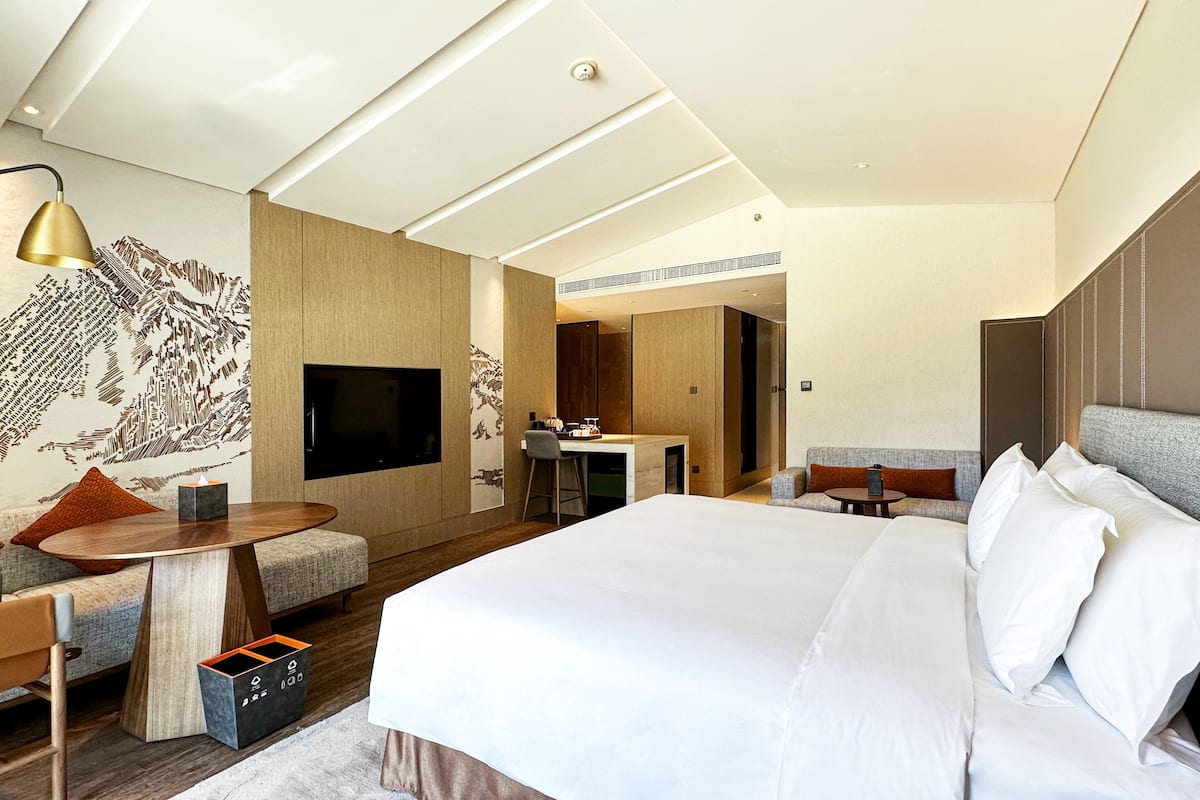 Habitación de hotel moderna con una cama grande, un escritorio, un televisor y una zona de estar con sofá y sillones.