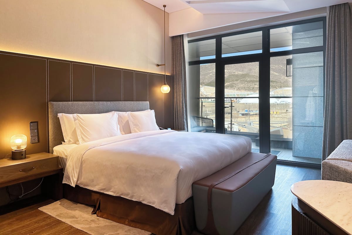 Ein modernes Hotelzimmer mit einem großen Bett, einem Nachttisch mit Lampe, einer Bank am Fußende des Bettes und einem großen Fenster mit Blick ins Freie.