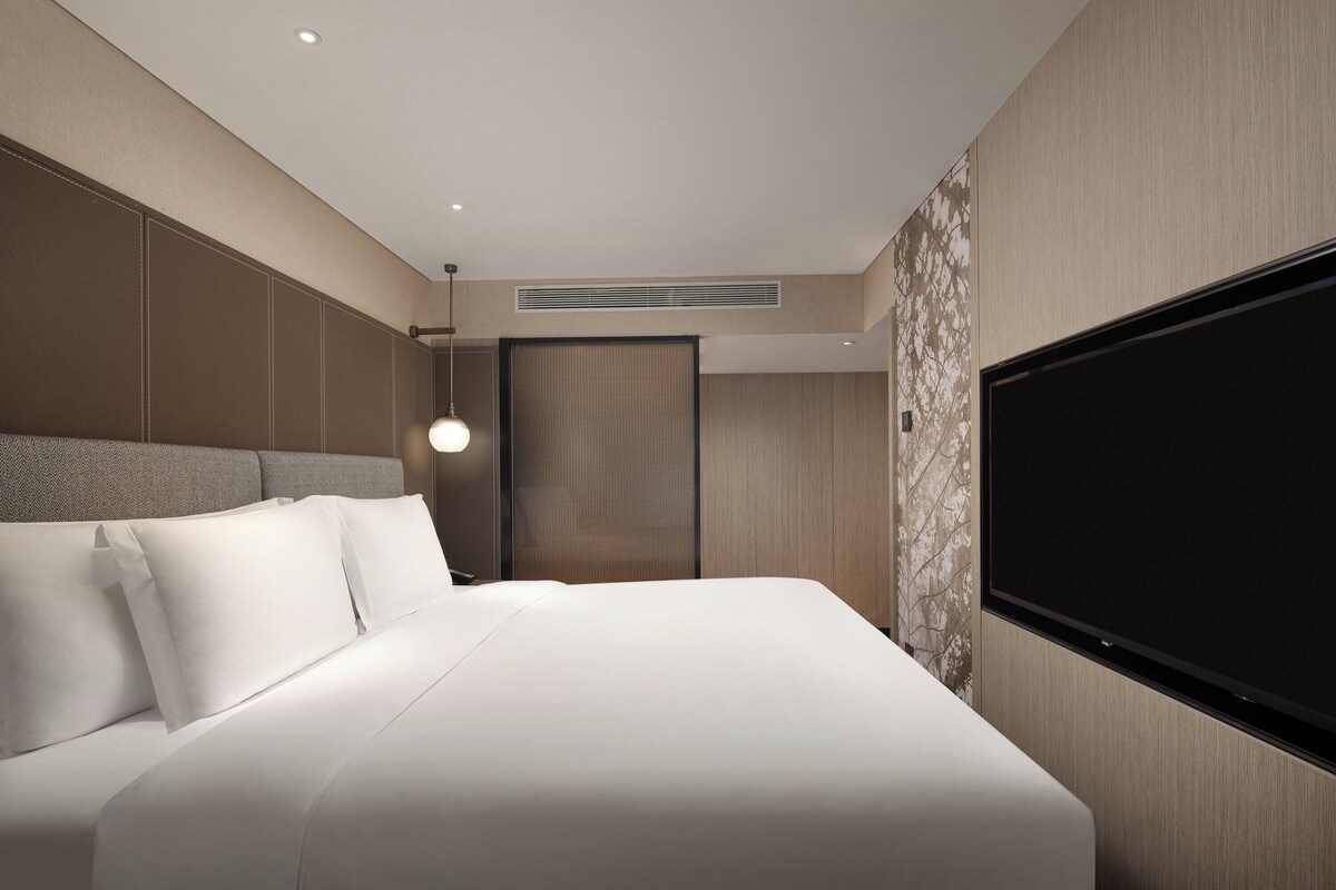A modern hotel room with a large bed, a flat-screen TV mounted on the wall, and a ceiling vent.