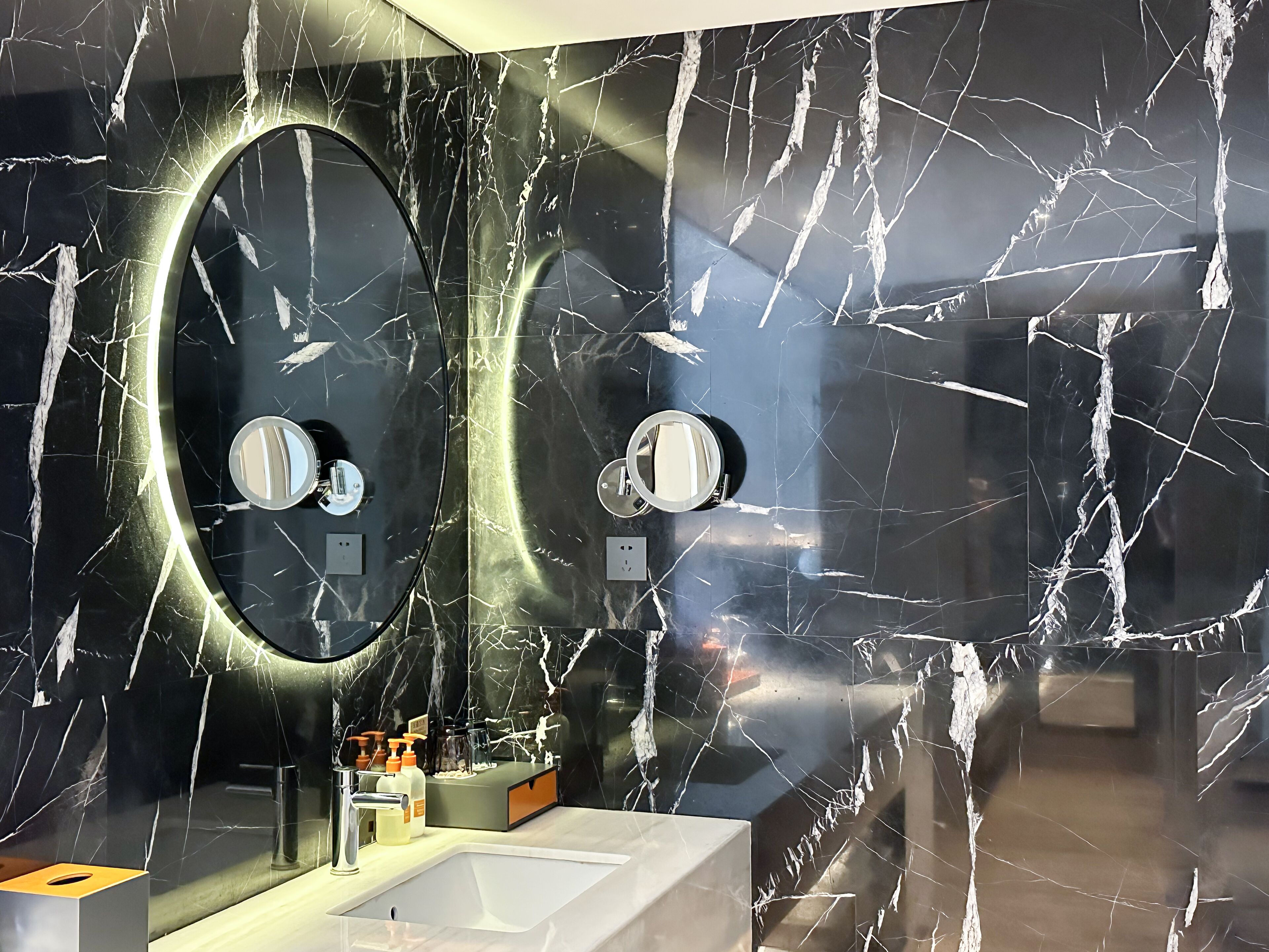 A bathroom with a black marble wall, two oval mirrors, a white sink, and a modern faucet.