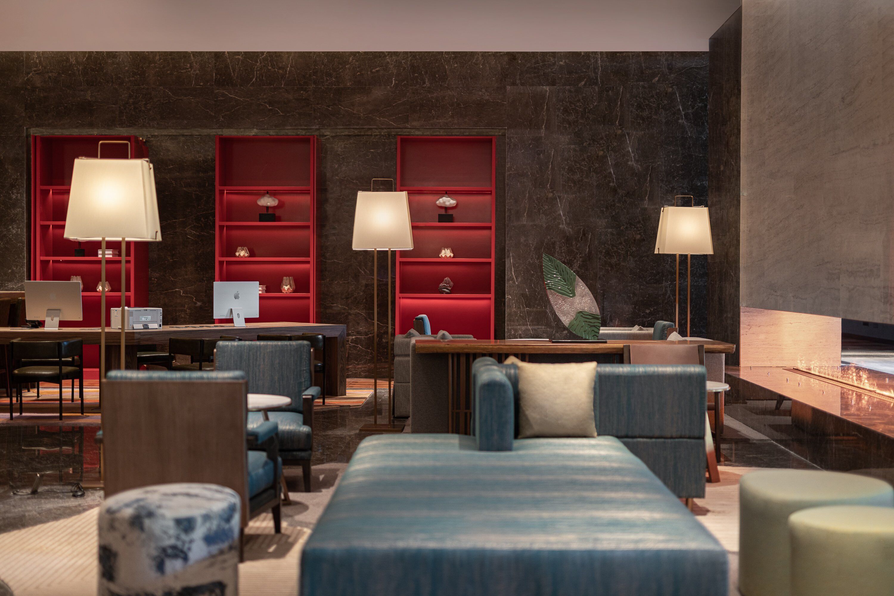 Une chambre d’hôtel moderne dotée d’un grand lit, d’un coin salon et d’un bureau avec des ordinateurs. On y trouve des étagères rouges ornées d’objets décoratifs et de lampes.