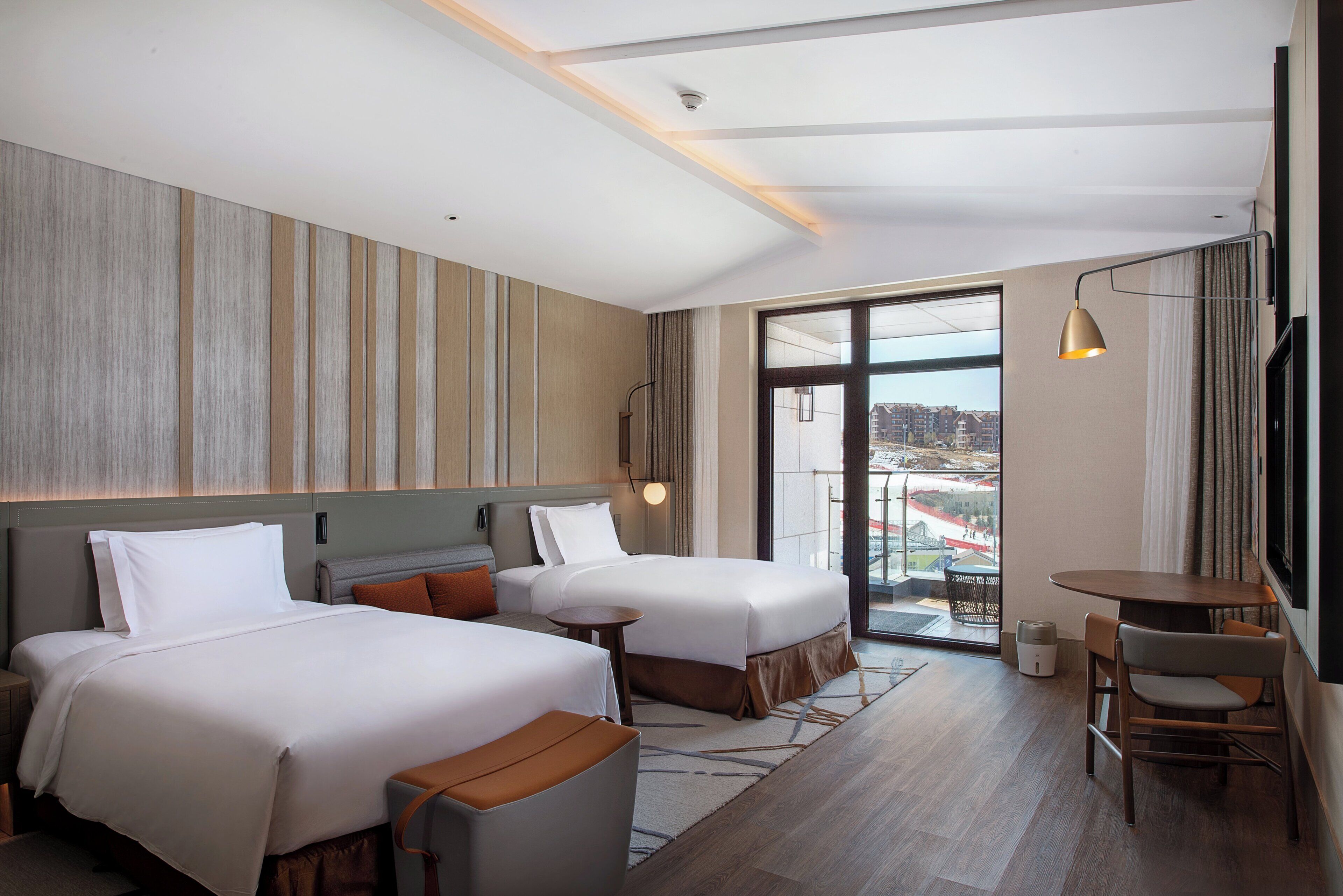 A hotel room with two beds, a small table, a chair, a balcony with a view, and a large window.