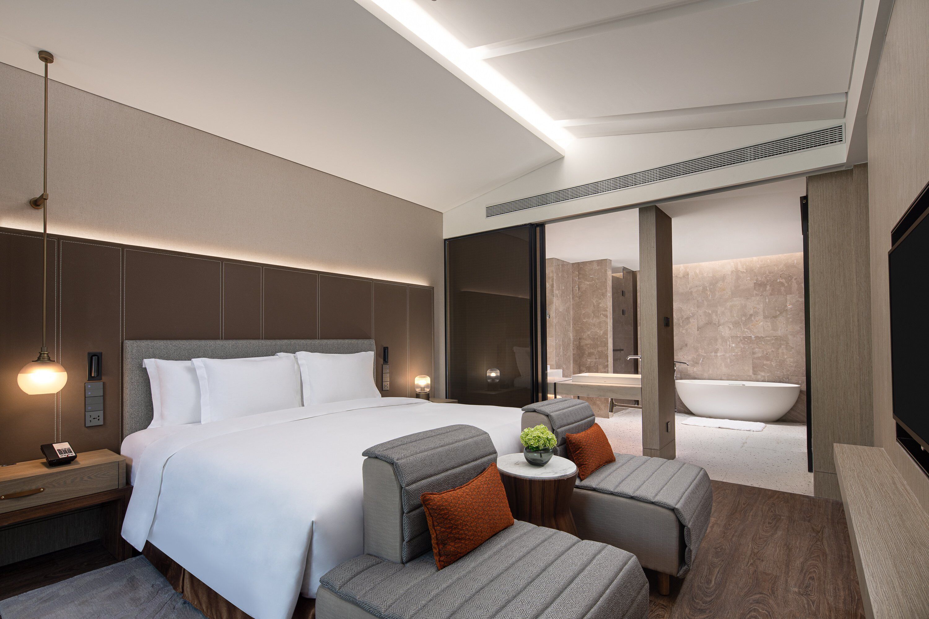 A modern hotel room with a large bed, two armchairs, a small table, a bedside lamp, a TV, and a bathroom with a bathtub and sink.