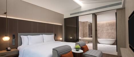 A modern hotel room with a large bed, two armchairs, a small table, a bedside lamp, a TV, and a bathroom with a bathtub and sink.