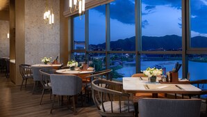 A restaurant with large windows offering a view of a river and mountains. Tables are set with chairs and centerpieces.