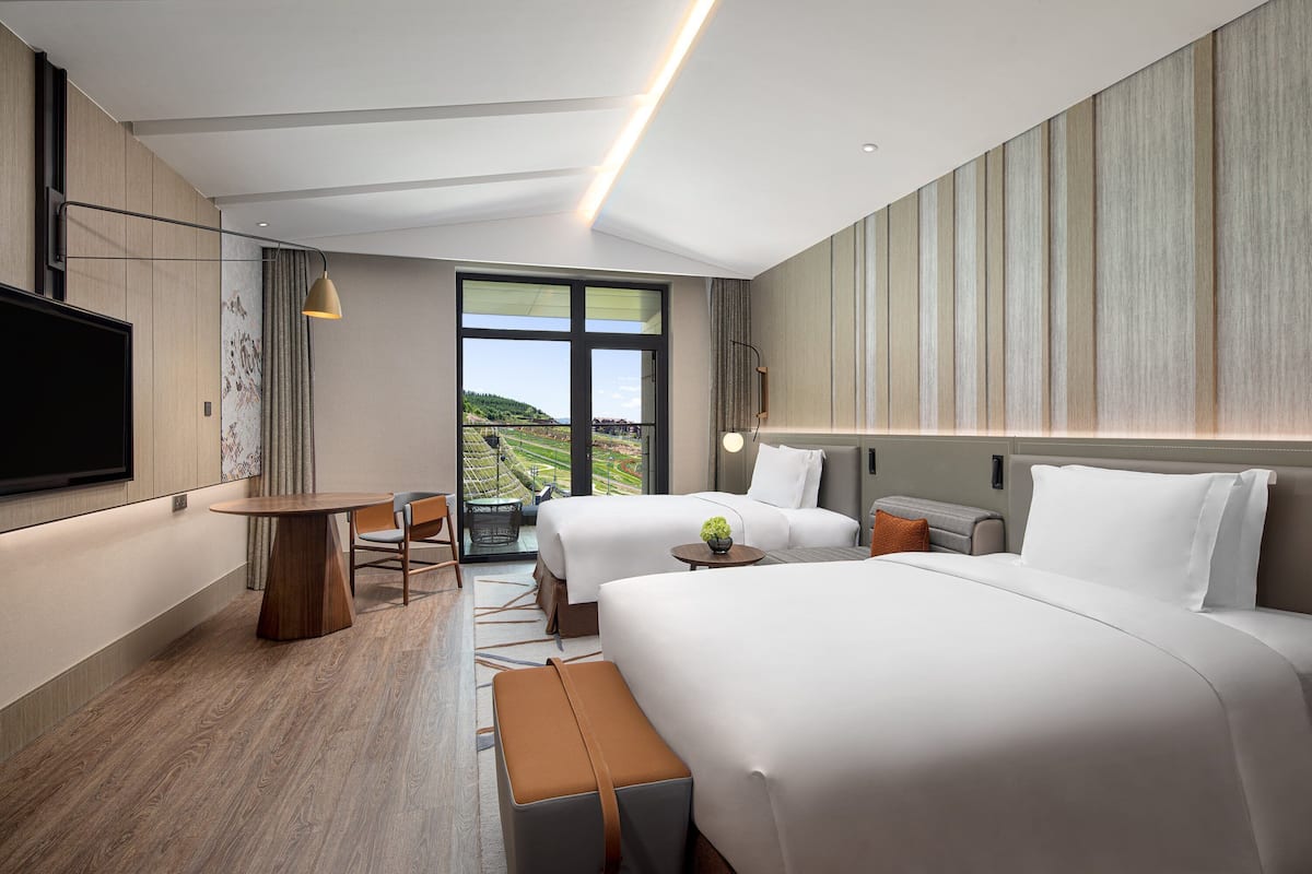 Ein modernes Hotelzimmer mit zwei Betten, einem Flachbildfernseher, einem Esstisch und Blick ins Freie.