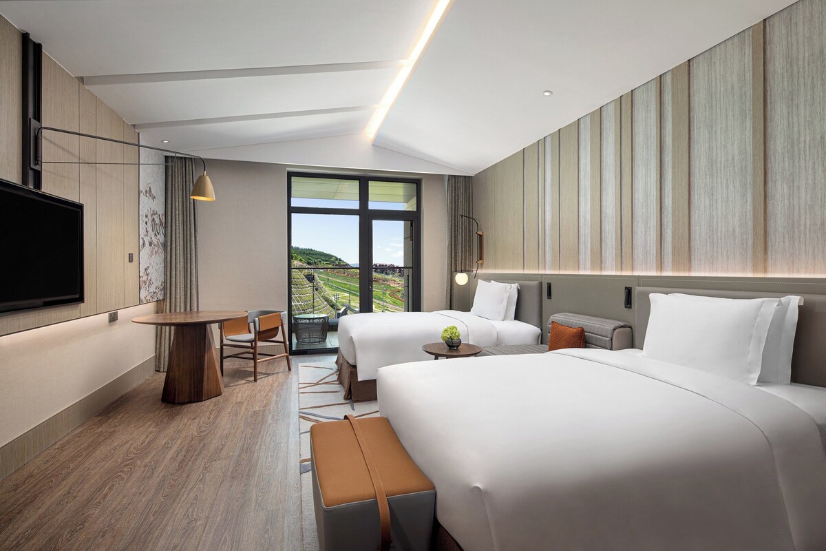 A modern hotel room with two beds, a flat-screen TV, a dining table, and a view of the outdoors.