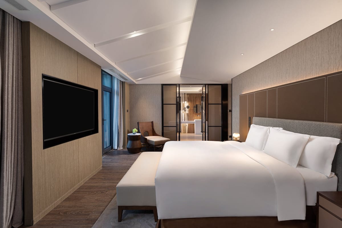 Ein modernes Hotelzimmer mit einem großen Bett, einem Flachbildfernseher, einer Sitzecke mit Stuhl und Tisch sowie Blick ins Badezimmer.