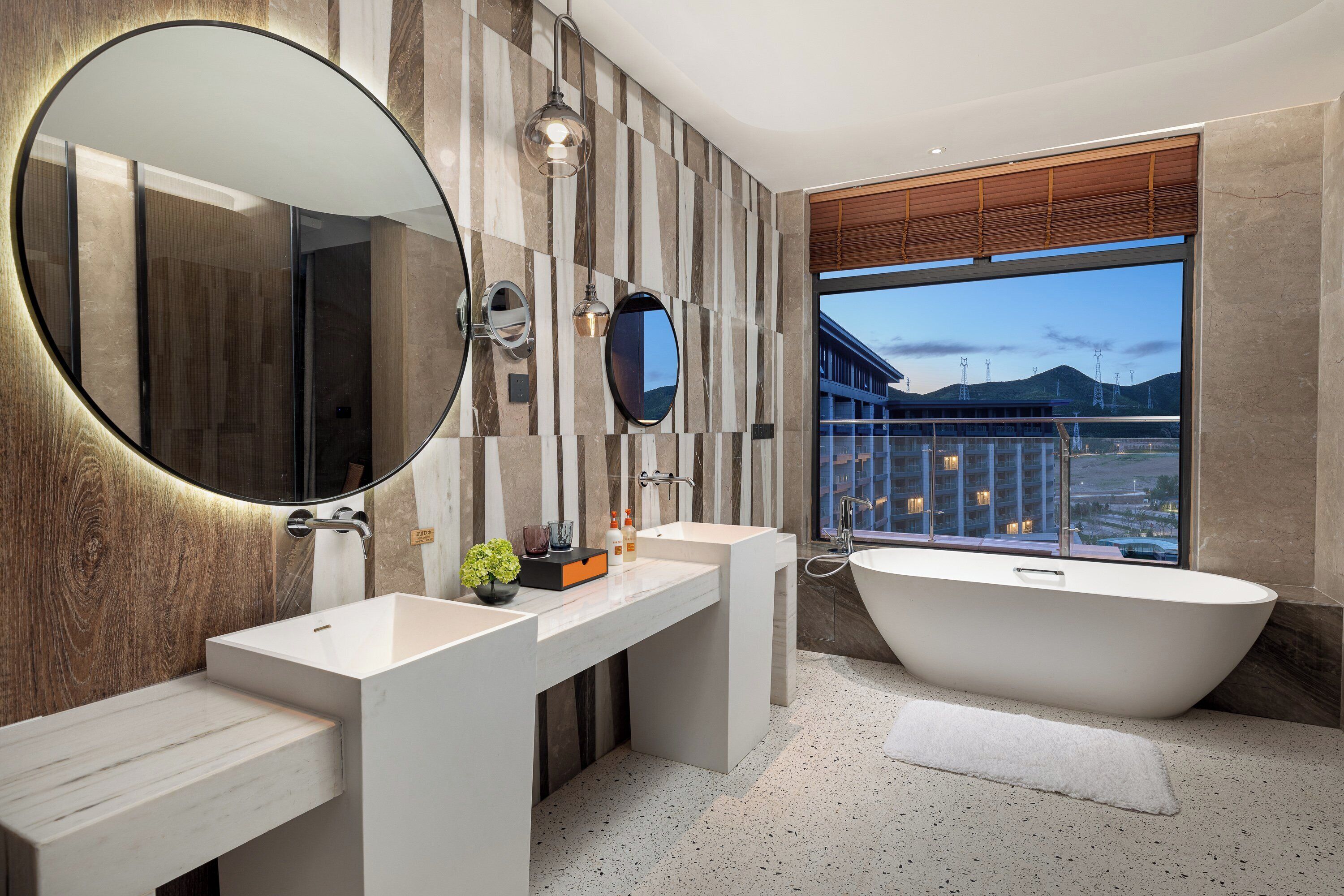 Une salle de bain moderne avec une grande baignoire, deux lavabos, un miroir et une vue sur un immeuble par la fenêtre.