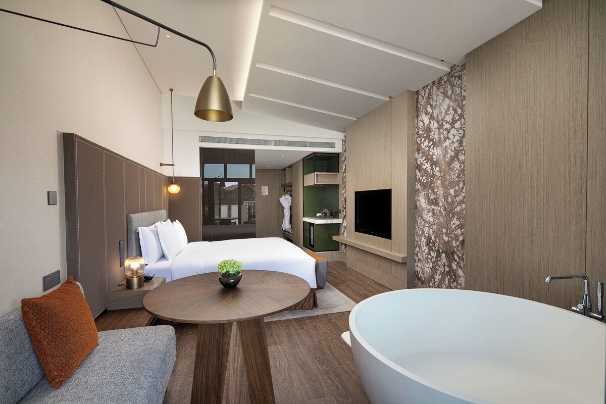 A modern hotel room with a large bed, a sitting area with a coffee table, a bathtub, and a view of the city.