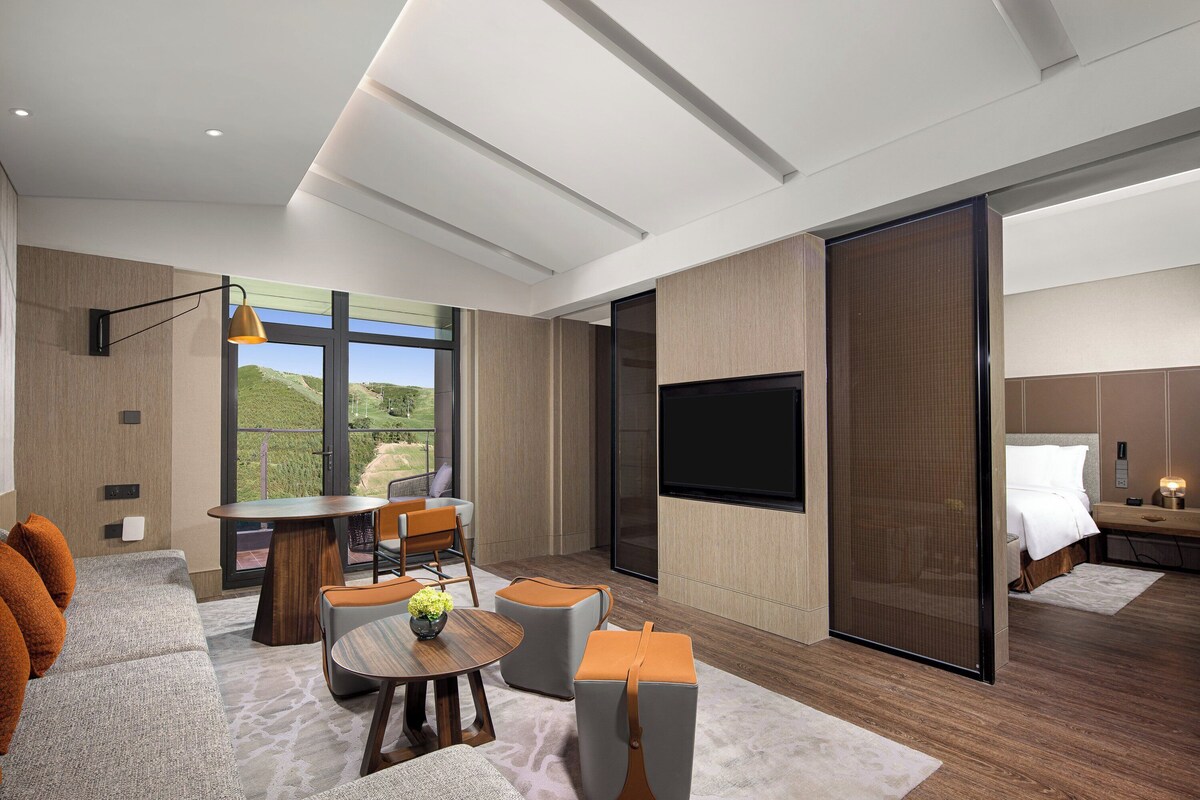 A modern hotel room with a large TV, a dining area with a table and chairs, a sofa, and a view of the outdoors.