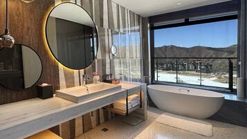 A modern bathroom with two sinks, a large bathtub, and a large window offering a view of a mountainous landscape.