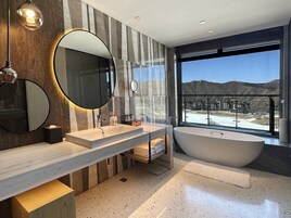 A modern bathroom with two sinks, a large bathtub, and a large window offering a view of a mountainous landscape.