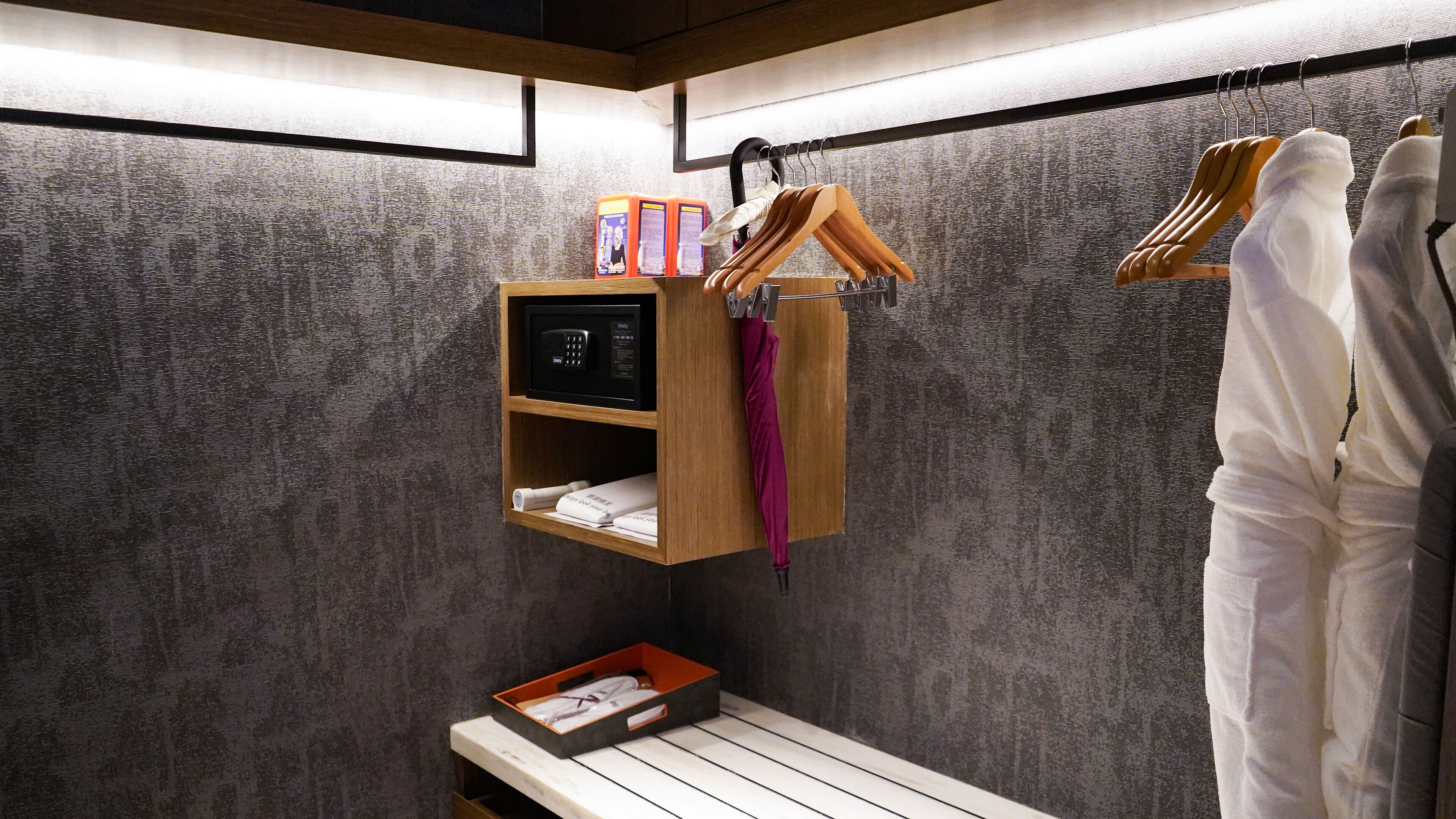 Habitación de hotel con un estante de madera que sostiene un dispositivo negro, una toalla morada y una bata blanca. Hay un banco de madera con una caja encima.