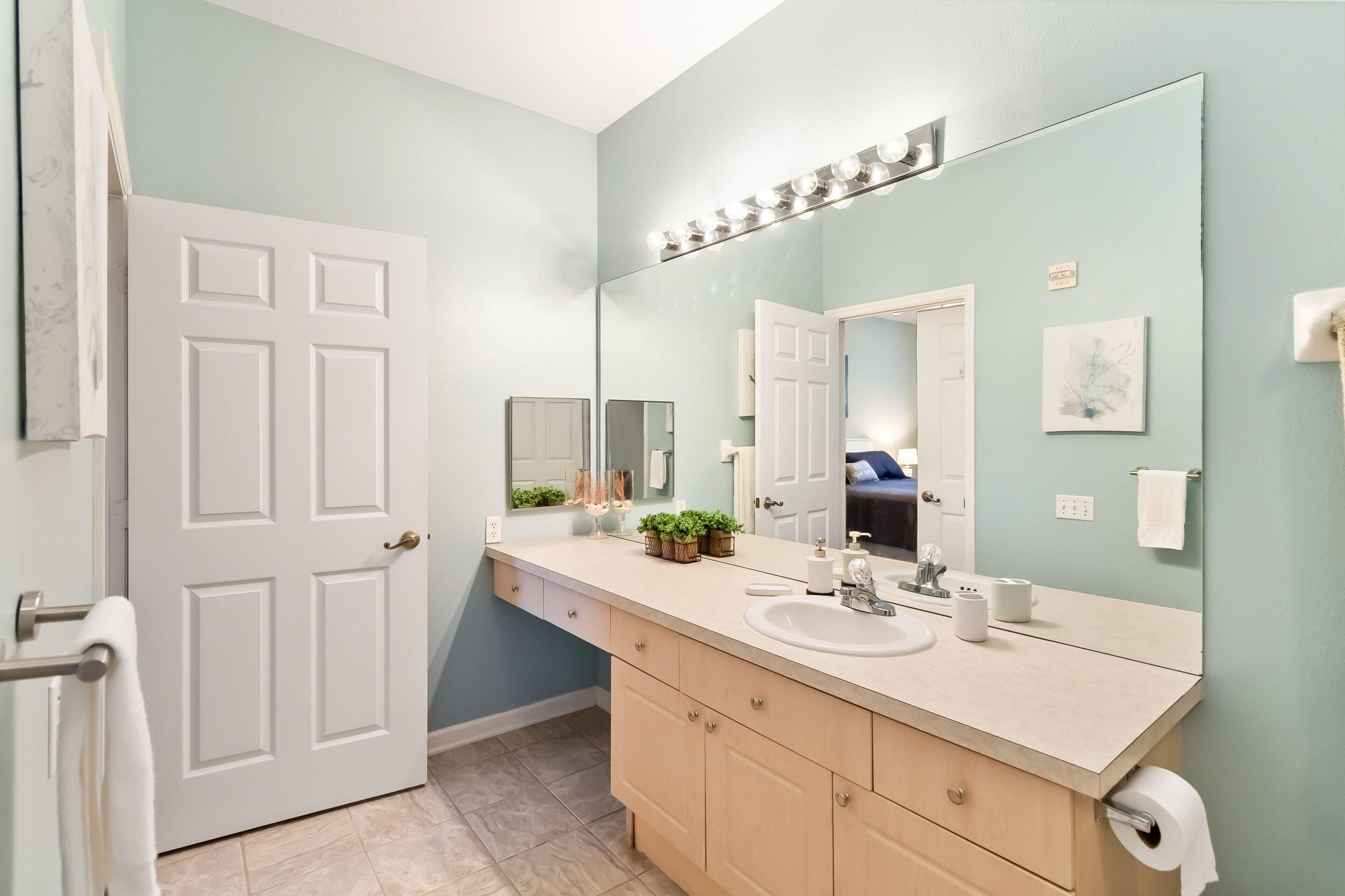 A bathroom with a white door, a large mirror with lights, a sink, and a toilet paper holder.