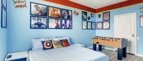 A bedroom with a bed, a foosball table, and superhero-themed wall art.