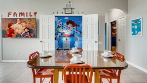 Ein Essbereich mit einem Glastisch und orangefarbenen Stühlen, einem Familienbild an der Wand und einem farbenfrohen Wandgemälde.