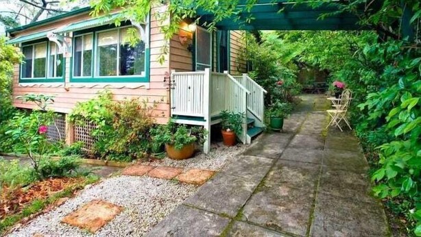 A small house with a porch, surrounded by lush greenery and a stone pathway. - Cherry Tree Cottage | Old-World Cottage, 3 Bedroom, A/C, Fireplace, Bath, Garden (Leura)