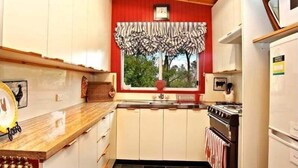 A compact kitchen with white cabinets, a window with striped curtains, a stove, and a refrigerator. - Cherry Tree Cottage | Old-World Cottage, 3 Bedroom, A/C, Fireplace, Bath, Garden (Leura)