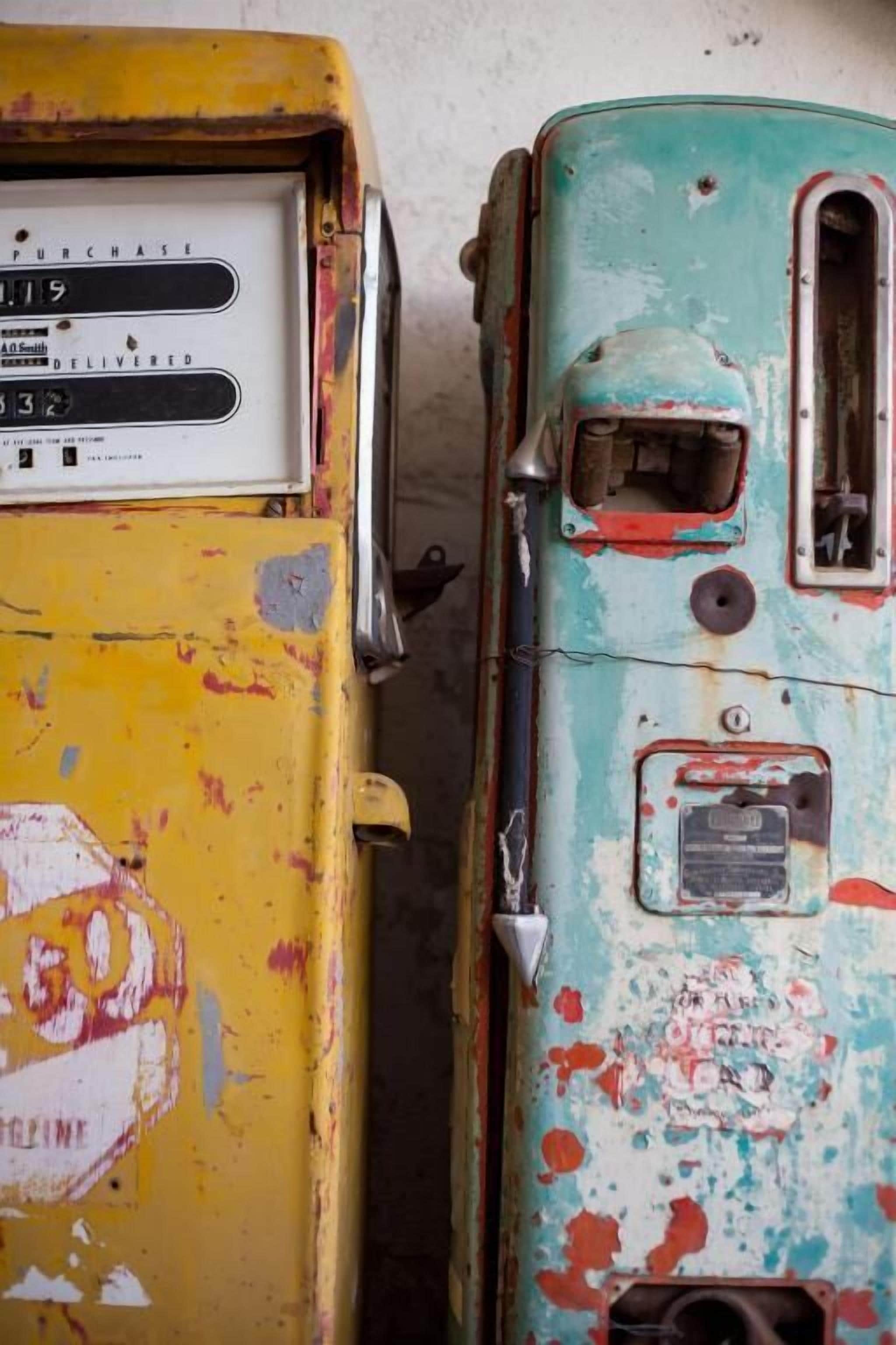 Two vintage vending machines with worn and peeling paint, one yellow and one teal, against a plain wall.